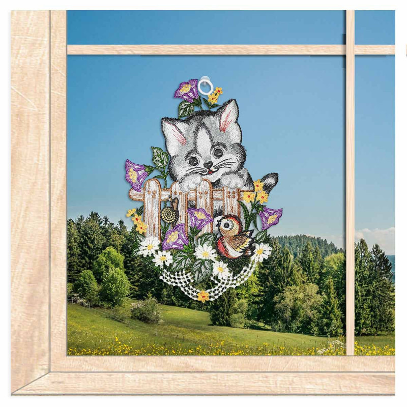 Gesticktes Fensterbild mit einer Katzen auf einem Holzzaun im idyllischen Frühlings-Ambiente am Fenster.