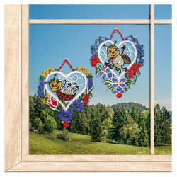 Zwei gestickte Fensterbilder in Herzform mit Marienkäfern vor einem Fenster mit Ausblick auf eine Berglandschaft.