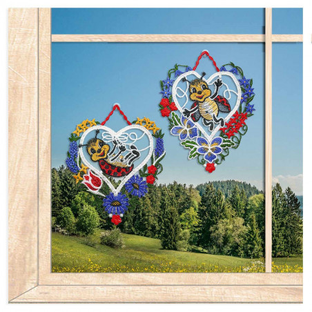 Zwei gestickte Fensterbilder in Herzform mit Marienkäfern vor einem Fenster mit Ausblick auf eine Berglandschaft.