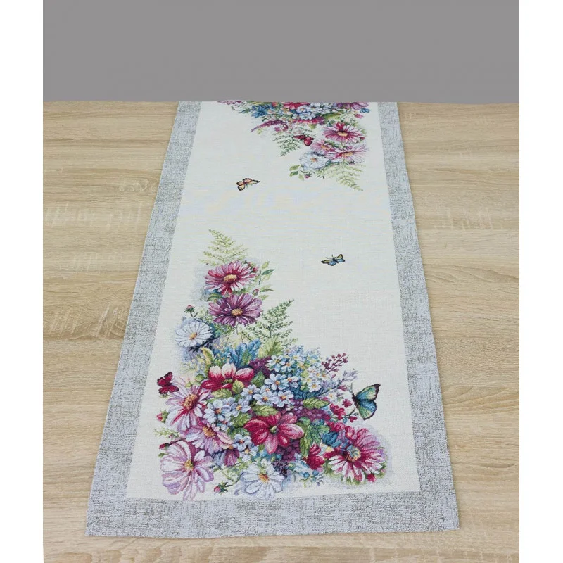 Der Tischläufer verfügt über einen cremefarbenen Grund mit einem markanten, grau melierten Randabschluss. Die Anordnung der Blumen sind in üppigen Eckmotiven zu sehen.