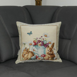 Auf einem grauen Sofa ist eine niedliches Osterkissen mit zwei Hasen und bunten Blumen in Gobelin-Webtechnick platziert.