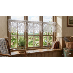 Die elegante weiße Spitzengardine Bella an einem sonnendurchflutetem Holzfenster.