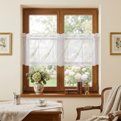 Die Plauener Spitze Kurzgardine Ayda in 30 cm Höhe mit moderne Ranken-Stickerei in Weiß-Hellbraun wurde hier an ein Holzfenster dekoriert. Davor steht ein antiker Tisch mit Stuhl und schöner Dekoration.