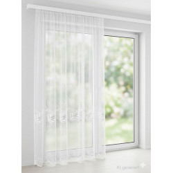 Bodenlanger, halbtransparenter Store in Weiß mit filigraner Plauener Spitze am Saum vor einer großen Fensterfront.