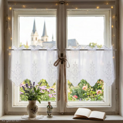 Weiße Kurzgardine aus Plauener Spitze mit detailreicher Stickerei am Fenster im Zimmer mit Blick ins Grüne als Heimtextilien.